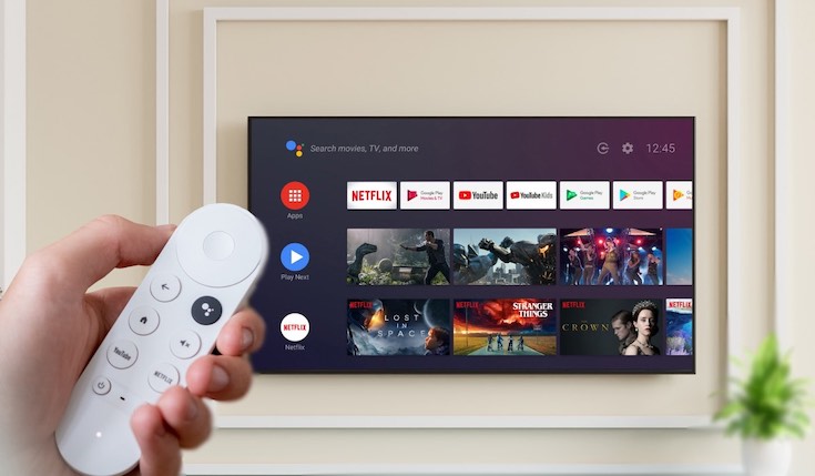A hand holding a Google Chromecast remote in the bottom-left corner. The remote is used to operate a Smart TV hanging on a wall. The Smart TV has a Google TV interface on the screen.