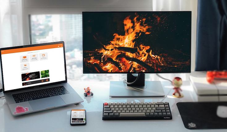 A desk with a monitor, a keyboard, a laptop, an iPhone and a mousepad. The monitor displays an image of flames on dark background. The laptop and the iPhone display a Mirror For Fire TV AirBeamTV app interface