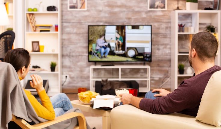 Two people in armchairs are watching TV. There are two shelving systems on each side of the TV