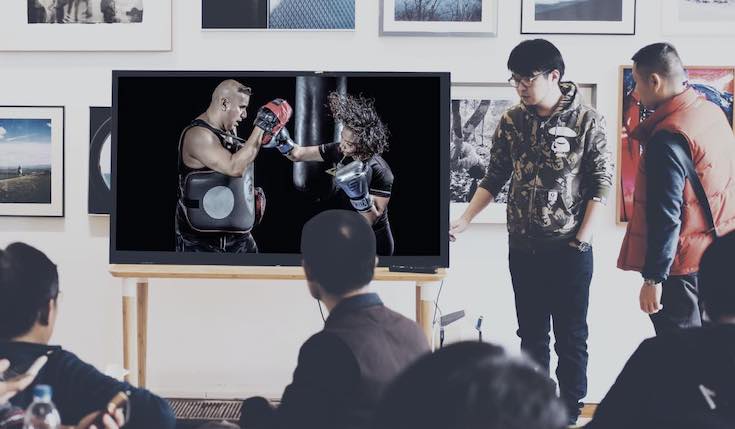 Two people on a stage next to a TV displaying a boxing match. There are several people watching the TV
