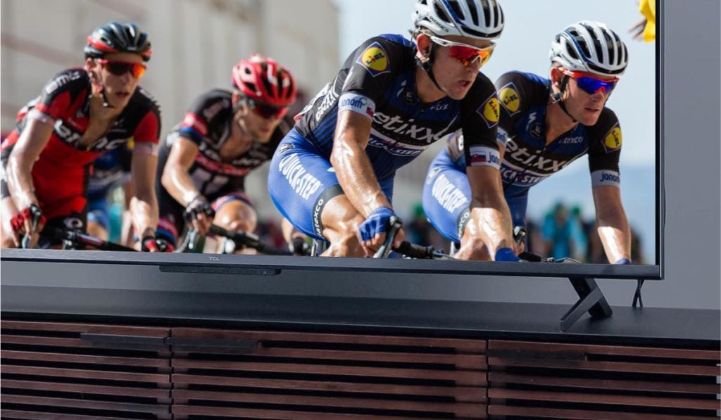 A TCL TV on a black TV drawer. The TV is displaying an image of four bike racers.