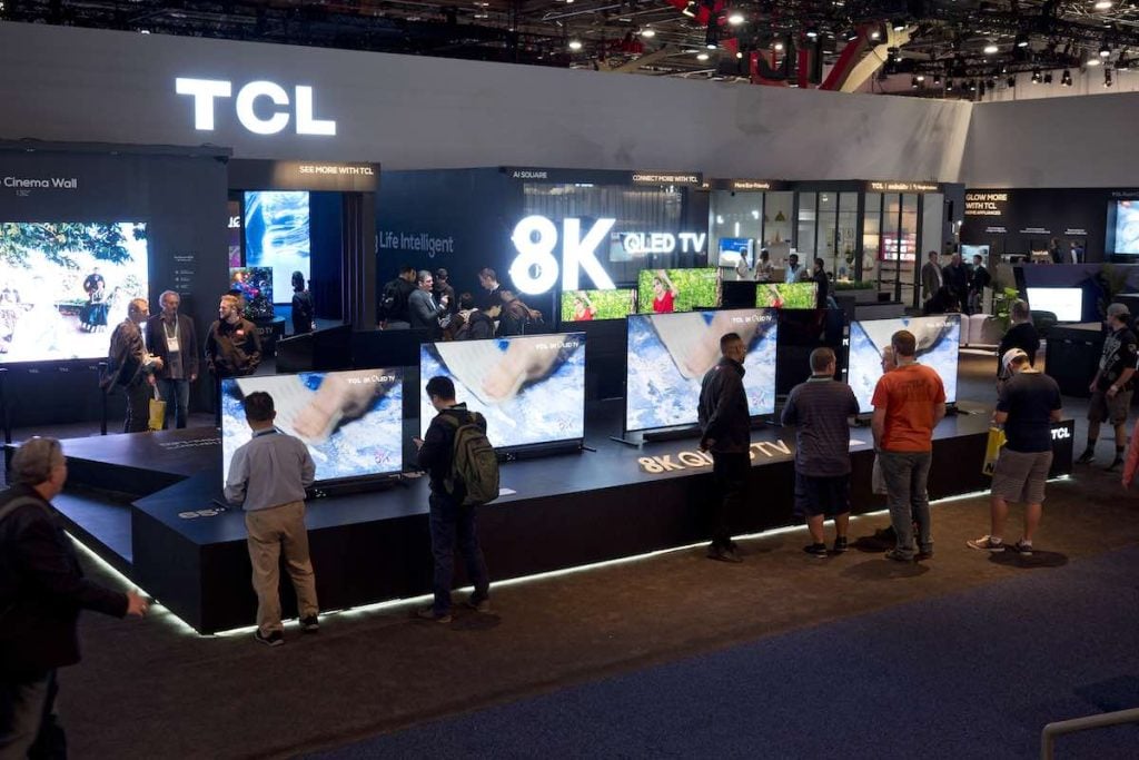 A large display containing several TCL TV units with large signs saying '8K' and 'TCL'. There are several people looking at the tVs.