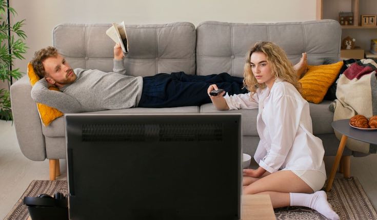 a man laying down on a couch and a woman sitting on the floor. The man is reading a newspaper while a woman is holding a remote and watching a TV screen.