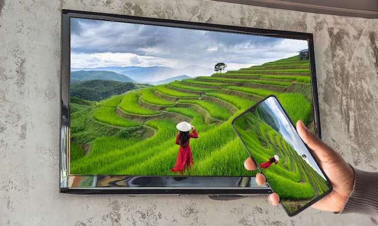 a hand holding a smartphone. The smartphone is casting an image of farmers tending to a rice field to a TV. The Tv is hanging on a wall