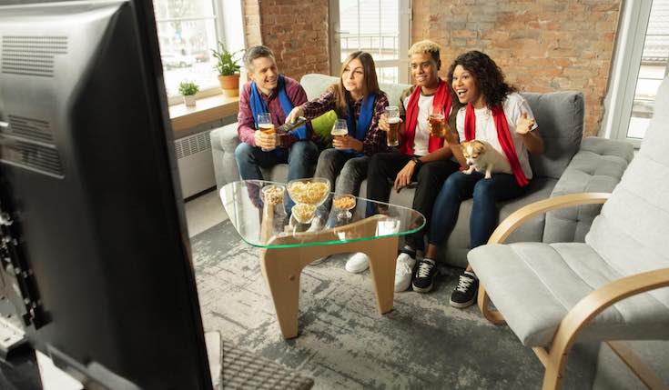 a group of friends sit on a couch watching TV. There's a chair next to them and a table with snacks and drinks in front of them