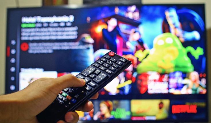 a hand holding a TV remote and pointing it at a TV that displays a streaming app interface