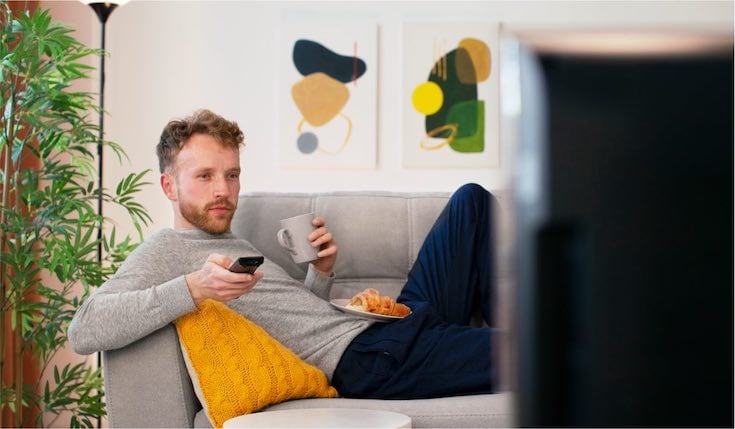 A man sitting comfortably on a couch with a cup and a remote in his hands watching tV