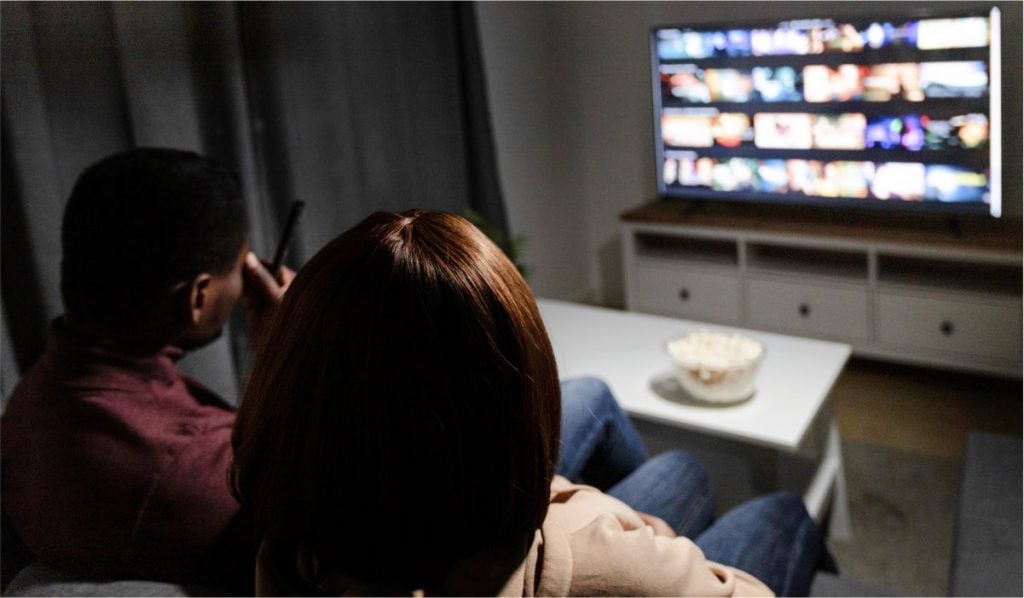 Two people sitting on a couch watching TV. They are seen from the back