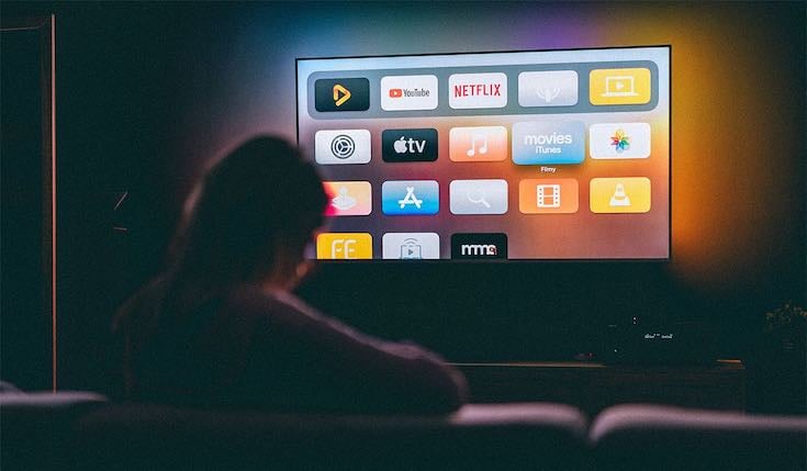 a person sitting in a dark room watching TV. The TV illuminates the room and has multiple app icons on the interface