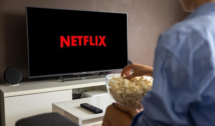 a person with a bowl of popcorn watching TV with Netflix logo on the screen. There is a coffee table in front of the TV with a remote control on it