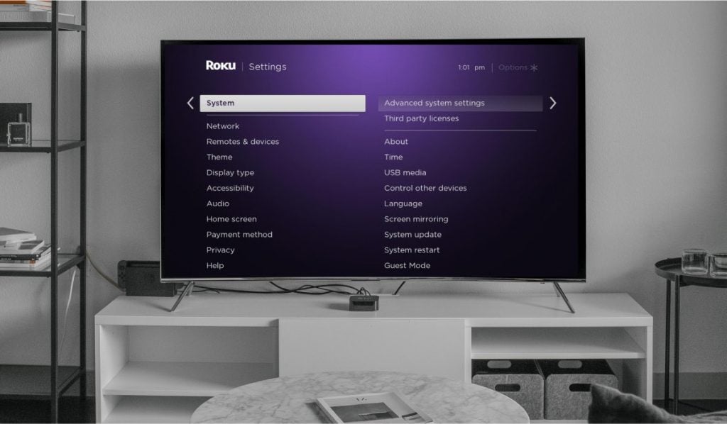 A Settings interface on a TCL Roku TV. The TV stands on a white drawer with shelves next to it