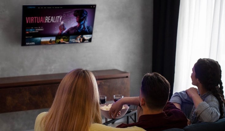 Three people sit on a couch watching TV. The TV is hanging on a wall