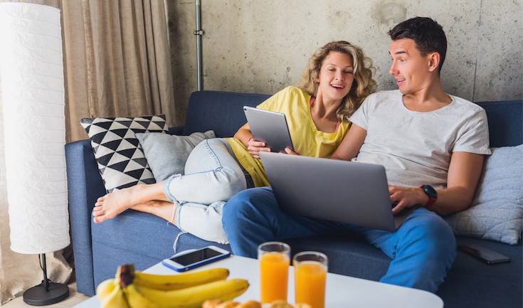 a man and a woman sit on a couch. The man uses a laptop and a woman uses a tablet. There is a table in front of them with bananas and orange juice