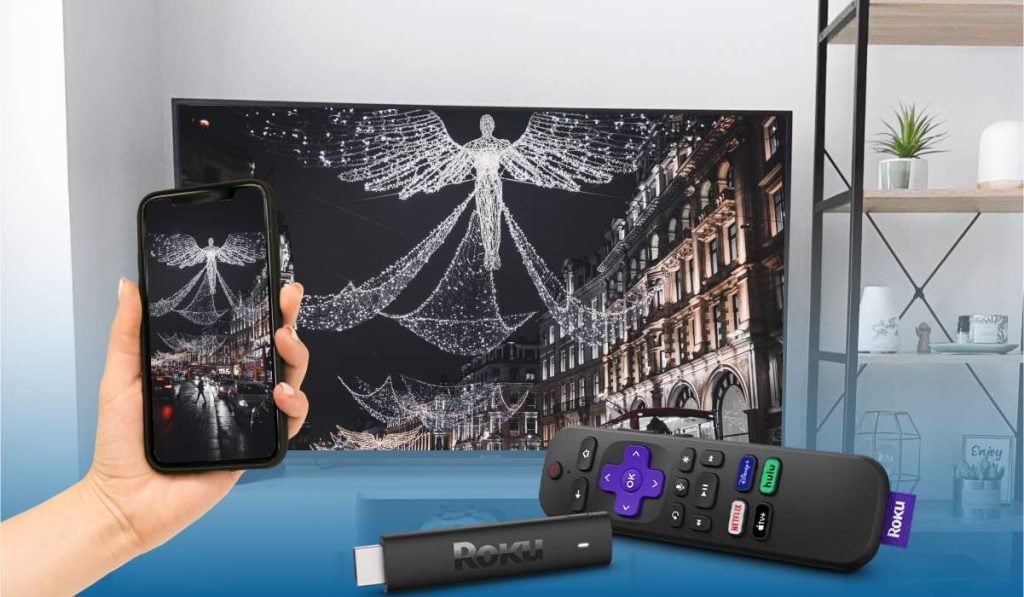 a hand holding an iphone that's casting na image of a christmas street to a TV. Roku device and remote