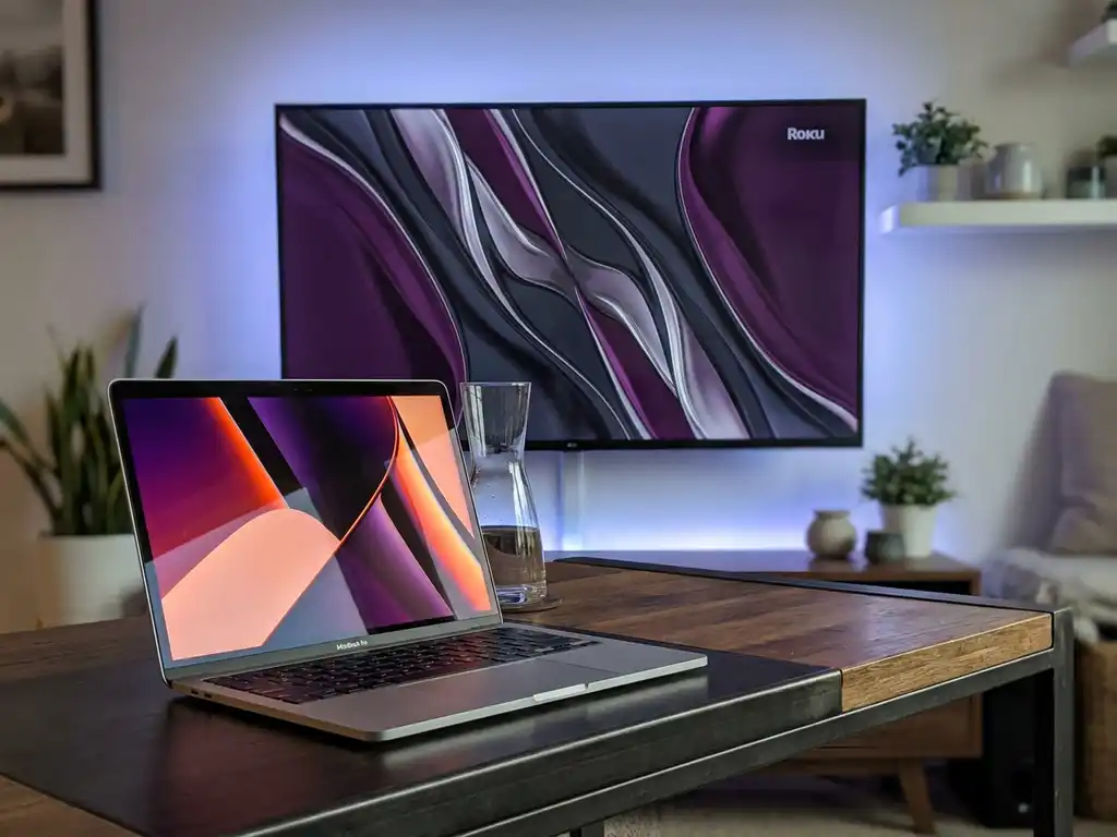 MacBook mirroring content to a Roku-connected TV on a modern desk with charcoal and purple ambient lighting.