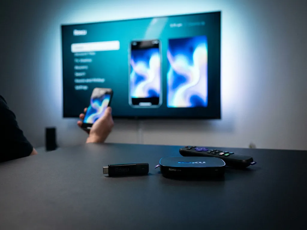 Roku Express and Roku Ultra streaming devices side by side, smartphone mirroring content to a glowing TV in the background.