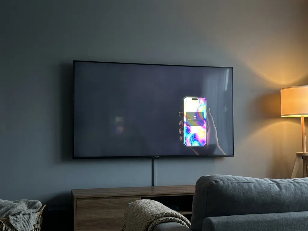 Sony Google TV mounted on a living room wall with a dim screen reflecting a glowing smartphone held nearby, warm ambient lighting.